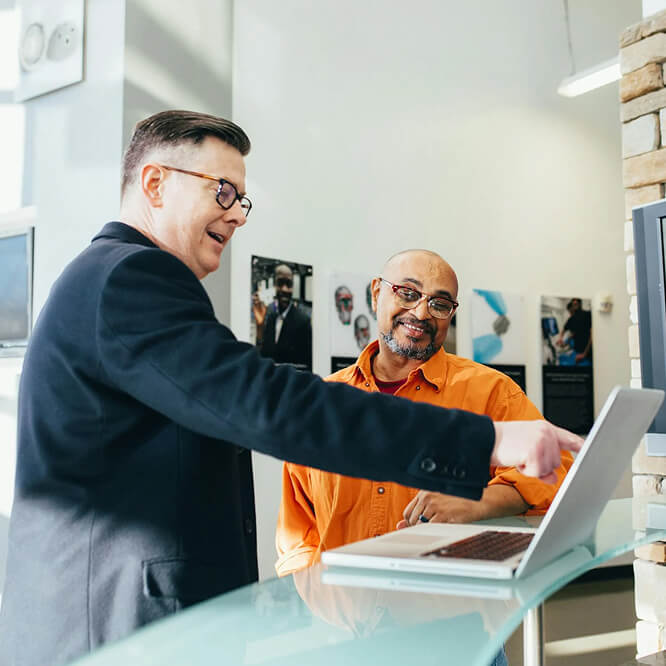 "Two men in conversation at a desk with a laptop. One man in a black jacket gestures towards the screen, while the other in an orange shirt smiles. The setting is a well-lit office with posters on the wall."