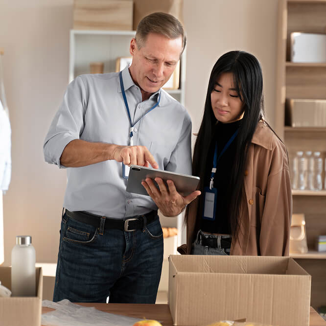 "Two people wearing name badges stand in an office setting, one holding a tablet while pointing at the screen. A cardboard box and assorted items are on the table in front of them."