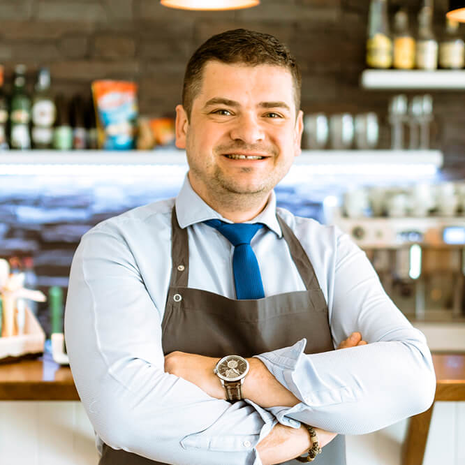 "A smiling man in a blue shirt and tie wearing a gray apron stands with arms crossed in a cafe. Shelves with bottles and a coffee machine are visible in the background."