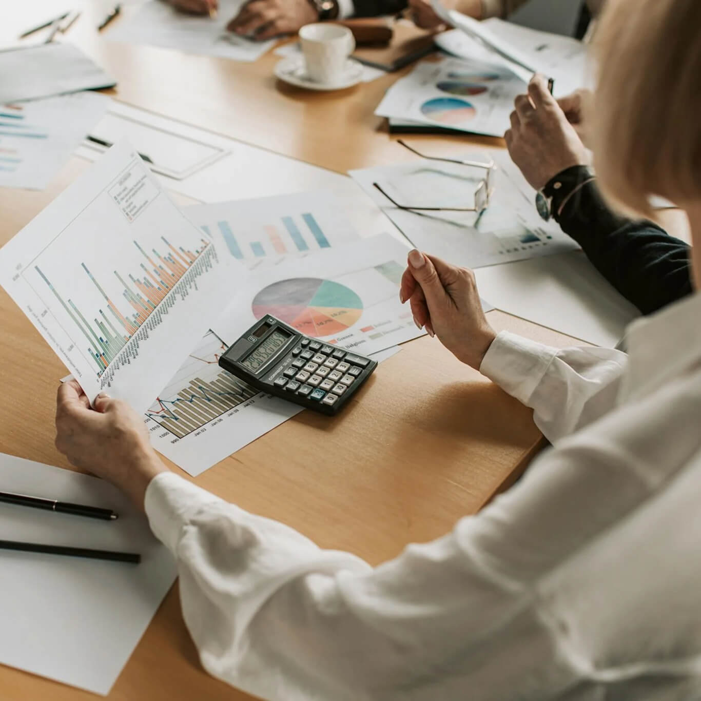  group of people sitting around a table reviewing various graphs and charts. One person holds a printed bar chart, and a calculator lies on top of the papers. A coffee cup is in the background.