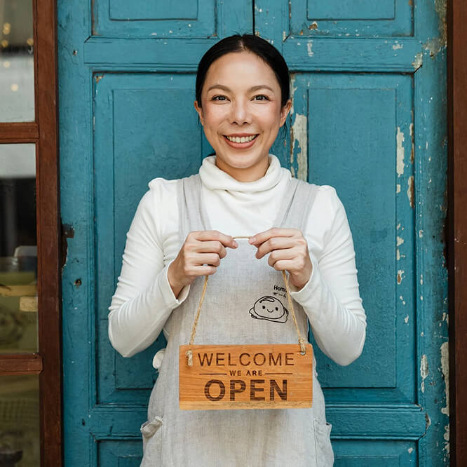 A smiling person in a white turtleneck and gray apron holds a wooden "Welcome We Are Open" sign. They stand in front of a rustic blue door.