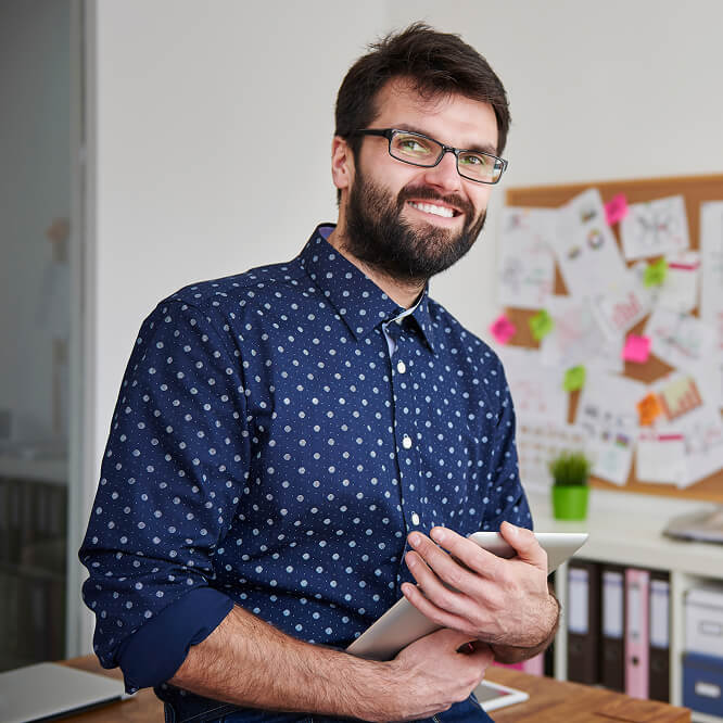 A man with glasses and a beard smiles while holding a tablet in an office. He wears a blue polka dot shirt. Behind him, a corkboard with papers and sticky notes, and a small plant on the desk, are visible.