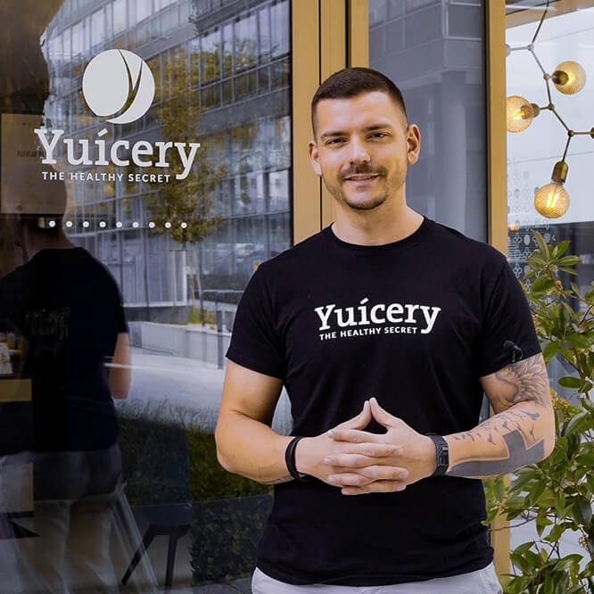 A man in a black "Yuicery - The Healthy Secret" shirt stands smiling in front of a shop window. The background features a modern building, a hanging light fixture, and greenery. Store hours and website are visible on the window.
