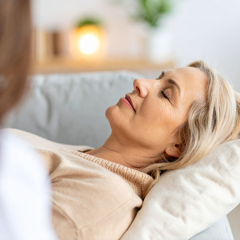 Frau mit geschlossenen Augen liegt entspannt auf einem Kissen auf dem Sofa.