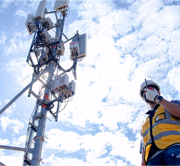 Field engineer wearing a safety vest and helmet, inspecting a cell tower with communication equipment against a cloudy sky.