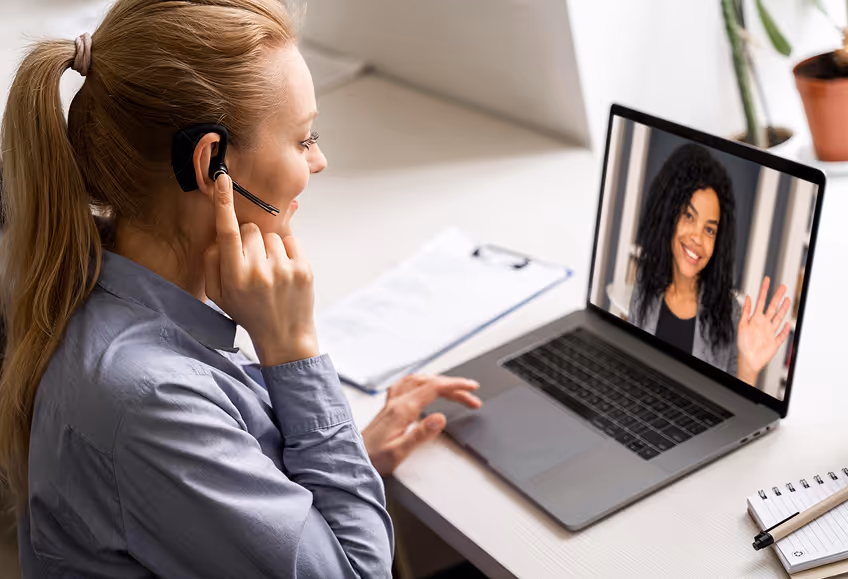 Female customer support agent wearing a headset having a video call with a smiling woman on a laptop screen.