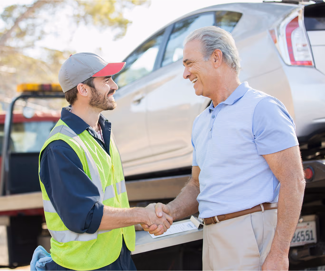 Tow truck worker in a reflective vest shaking hands with a smiling man in front of a car being towed.