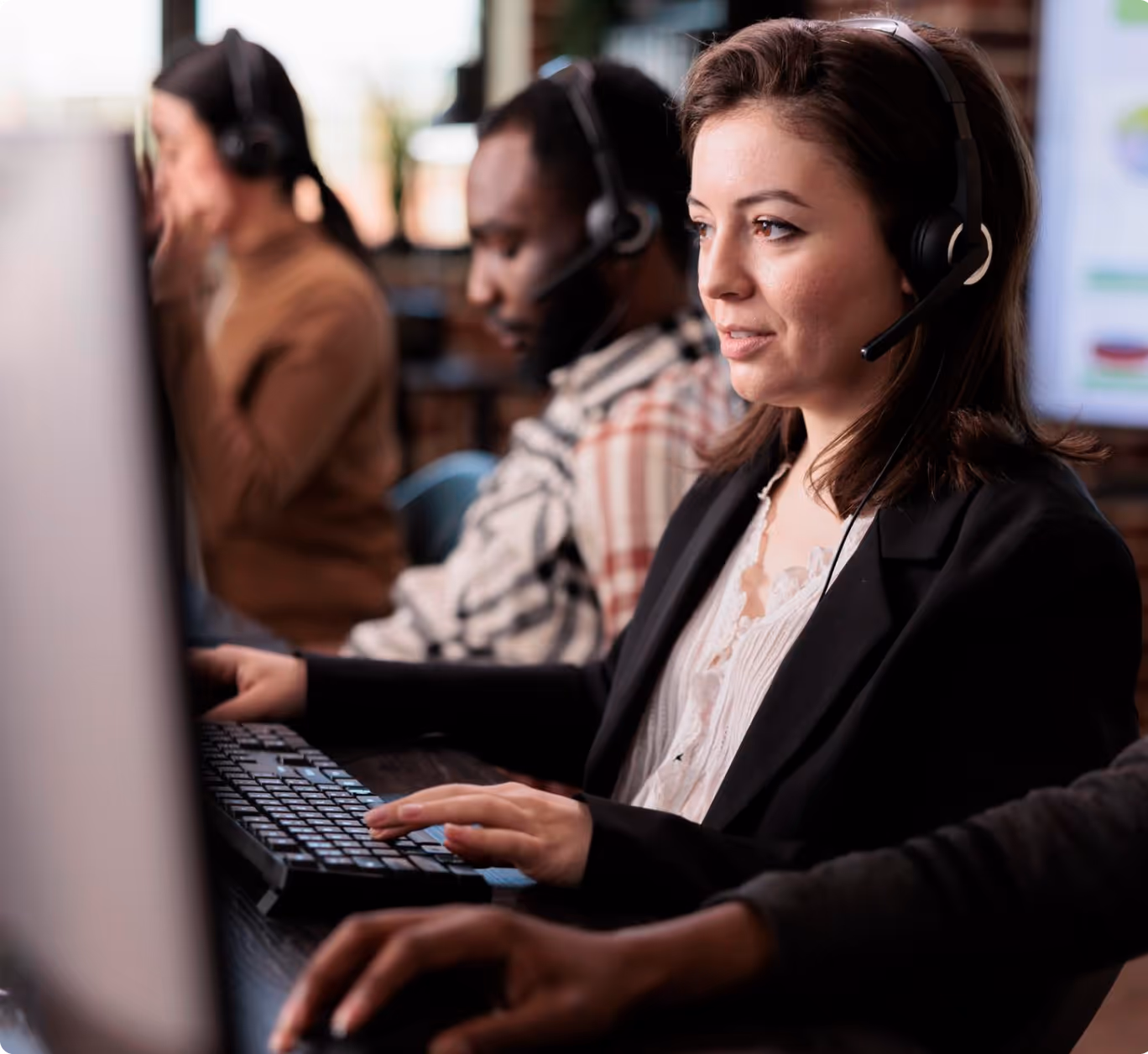 Focused female call center agent wearing a headset, typing on a keyboard with colleagues working in the background.