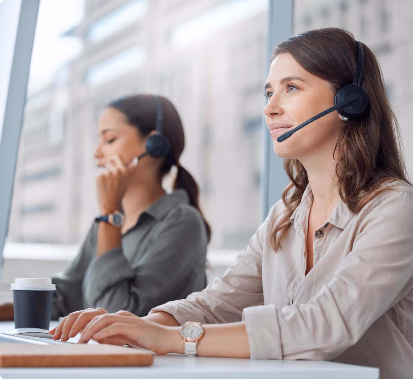 Two female customer service agents with headsets working at desks near large windows.