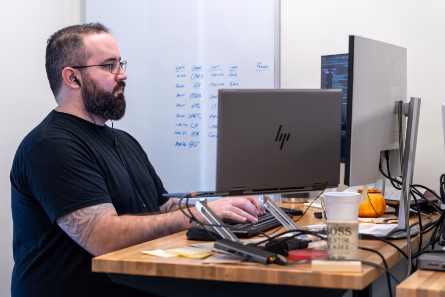 Man with glasses and a beard wearing a black t-shirt, working on a laptop and desktop computer at a desk with a coffee cup and office supplies.