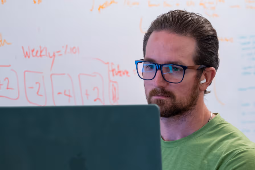 Man with glasses and a beard focused on a laptop screen, sitting in front of a whiteboard with red and orange handwritten notes.