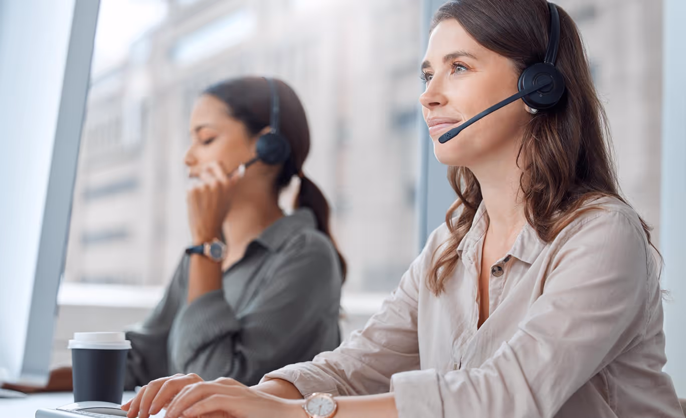 Two female call center agents wearing headsets, one typing and the other speaking on the phone in a bright office.