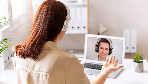Woman wearing headphones conducting a video call with a man on a laptop in a bright office setting.