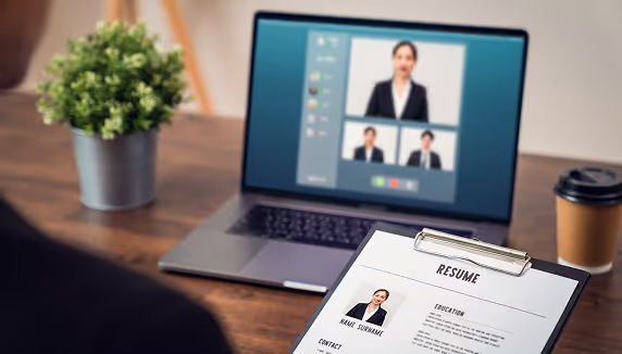 Person holding a resume on a clipboard with a laptop showing a video interview in the background on a wooden desk.