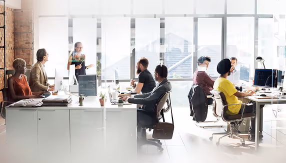 Group of diverse coworkers gathered in a modern office with large windows, some seated and others standing, engaged in discussion.