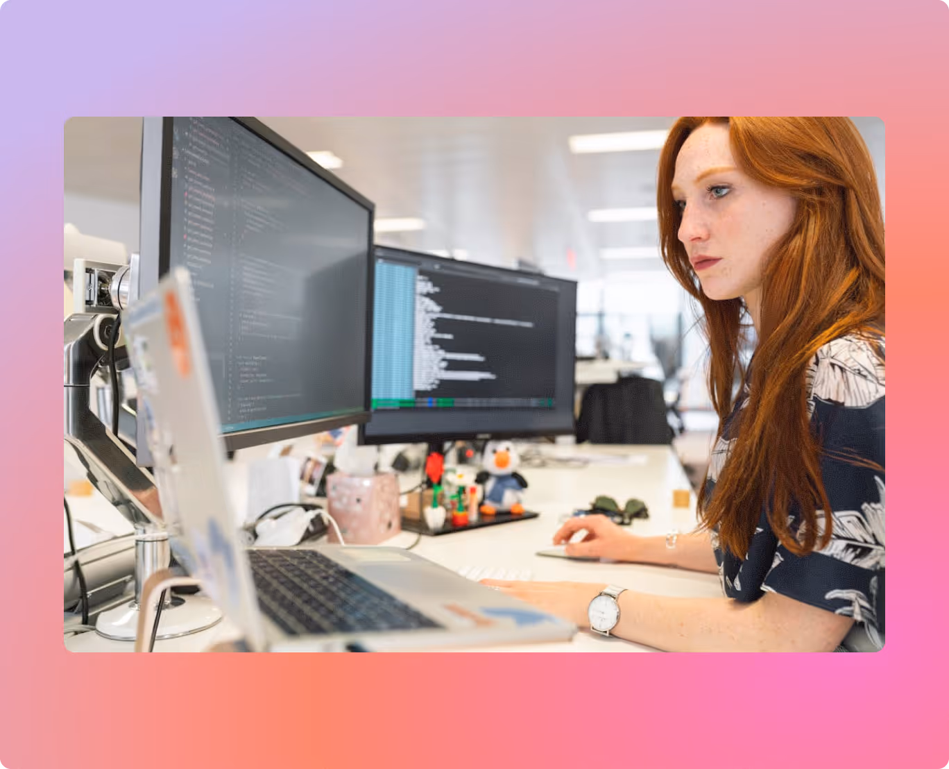 Red-haired woman focused on coding displayed on dual computer monitors at a bright office desk.