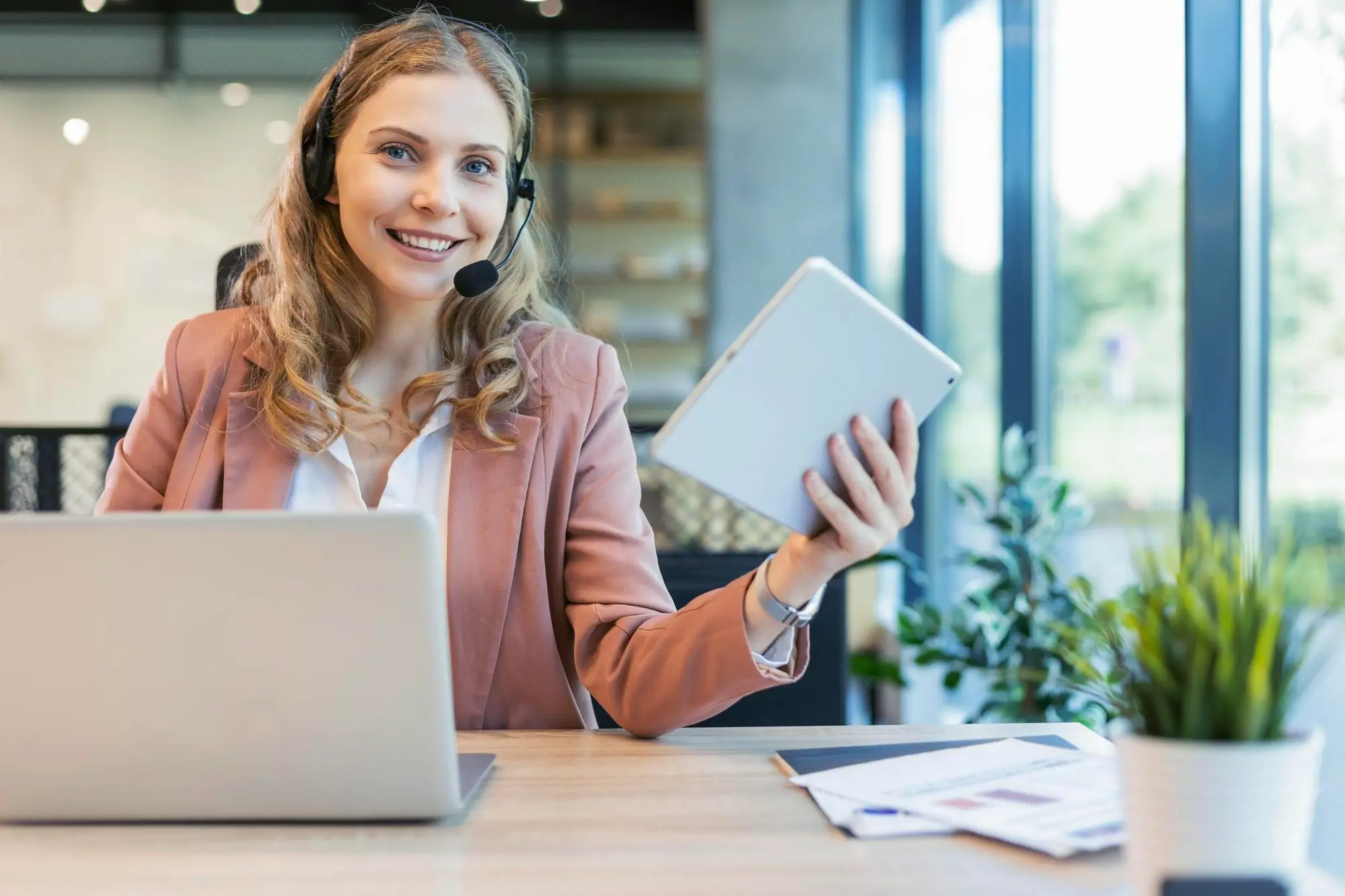 Smiling woman with headset holds tablet and works on laptop at desk in bright office.