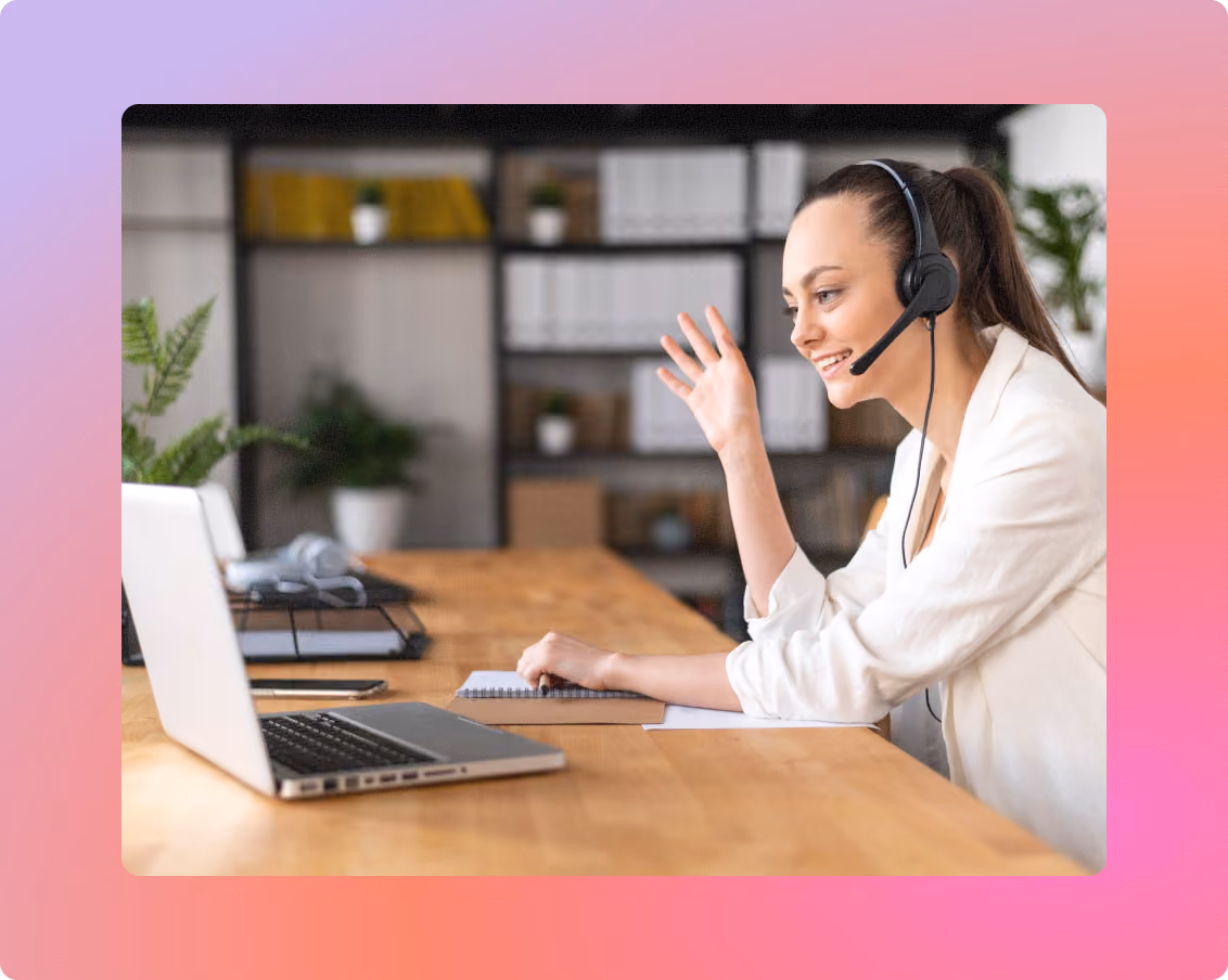 Woman wearing a headset smiling and waving at her laptop during a video call at an office desk.
