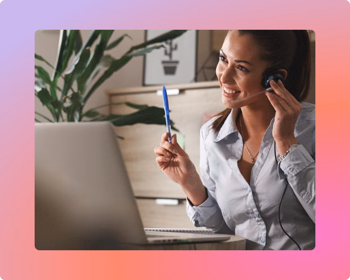 Woman wearing a headset smiling and holding a pen while talking to a customer in front of her laptop.
