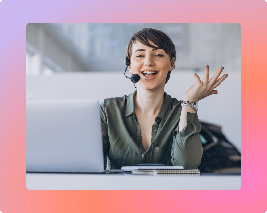 Woman wearing a headset laughing and gesturing with her hand while sitting at a laptop in a bright office.