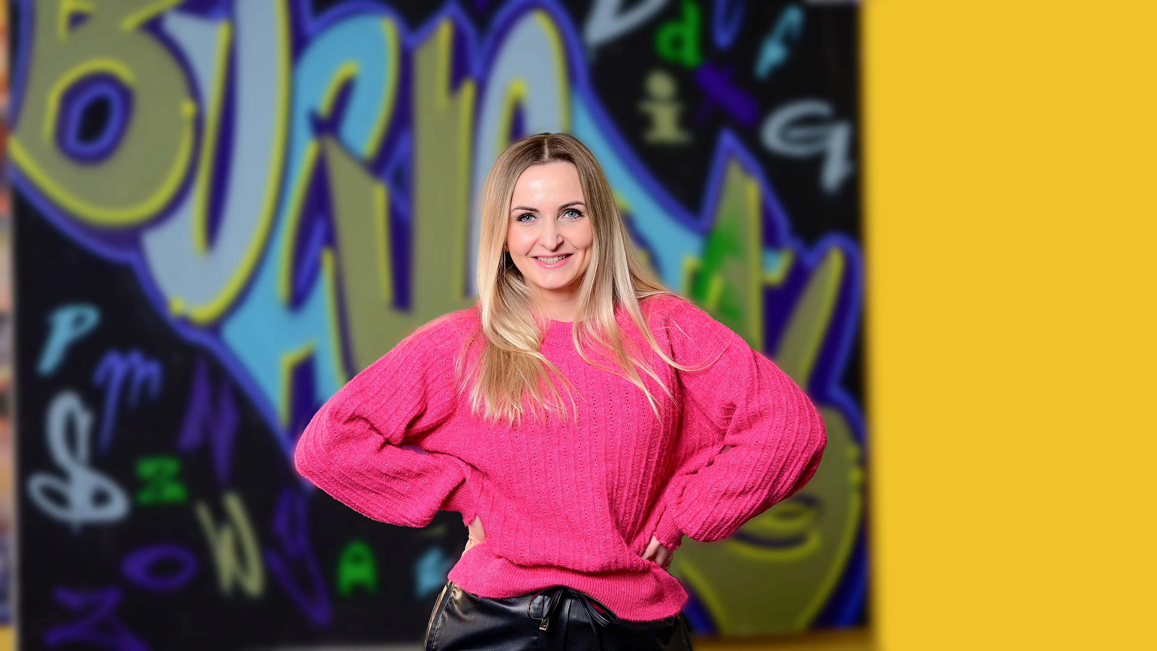 Lächelnde Frau mit langen blonden Haaren, trägt pinken Pullover und schwarze Hose vor bunter Graffiti-Wand.