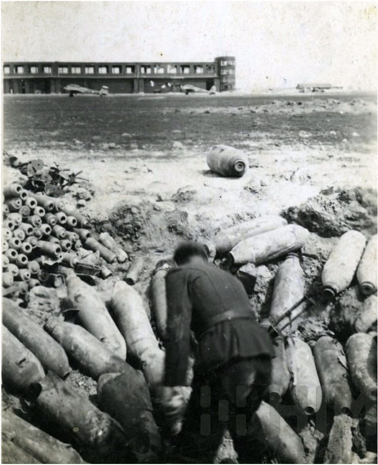 A tököli repülőtér bombázása után hatástalanított és összegyűjtött bombák, a háttérben a rombolt hangárokkal. (HTM)