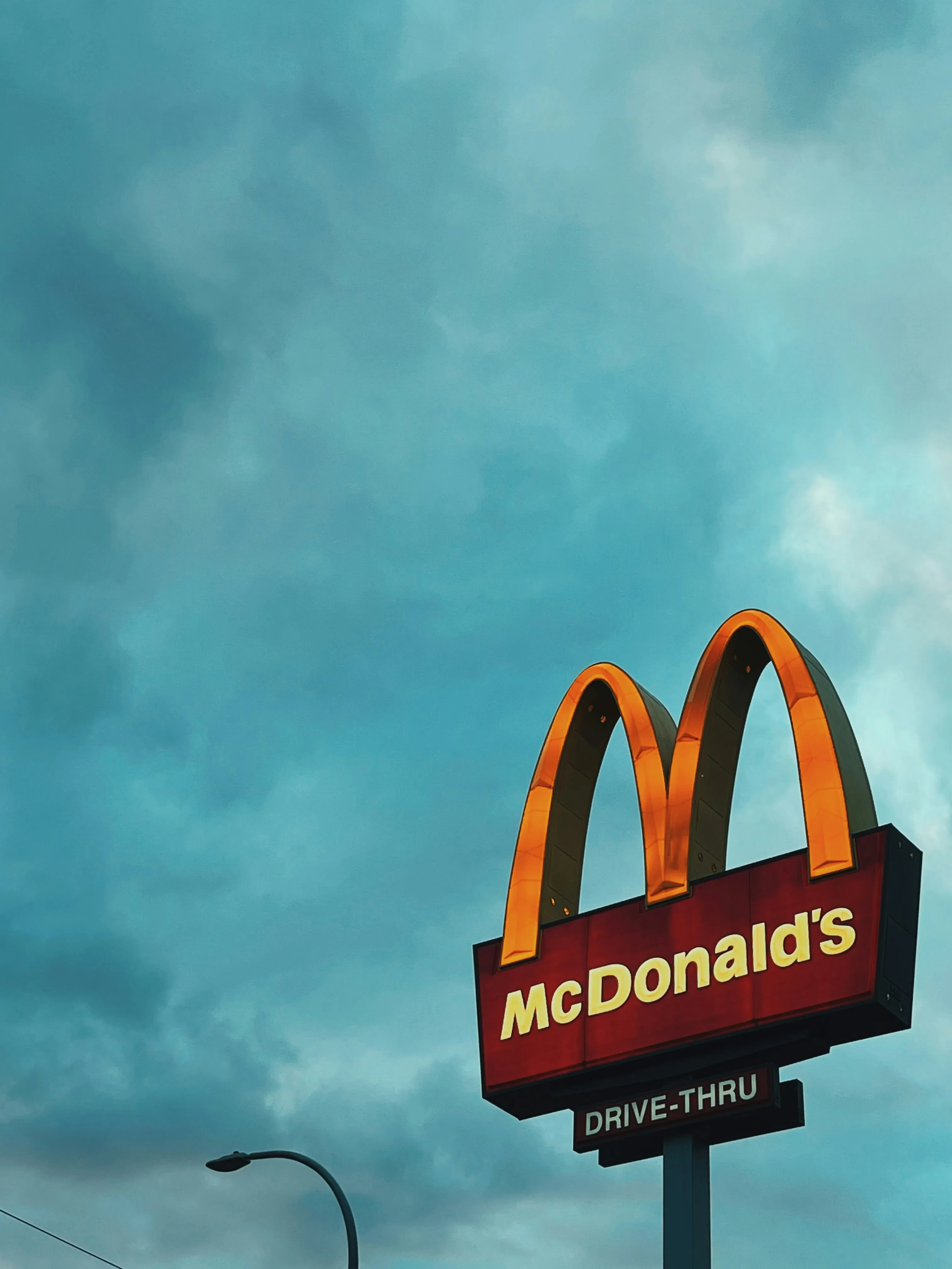 McDonald's sign with golden arches and 'Drive-Thru' on a pole against a cloudy blue sky.