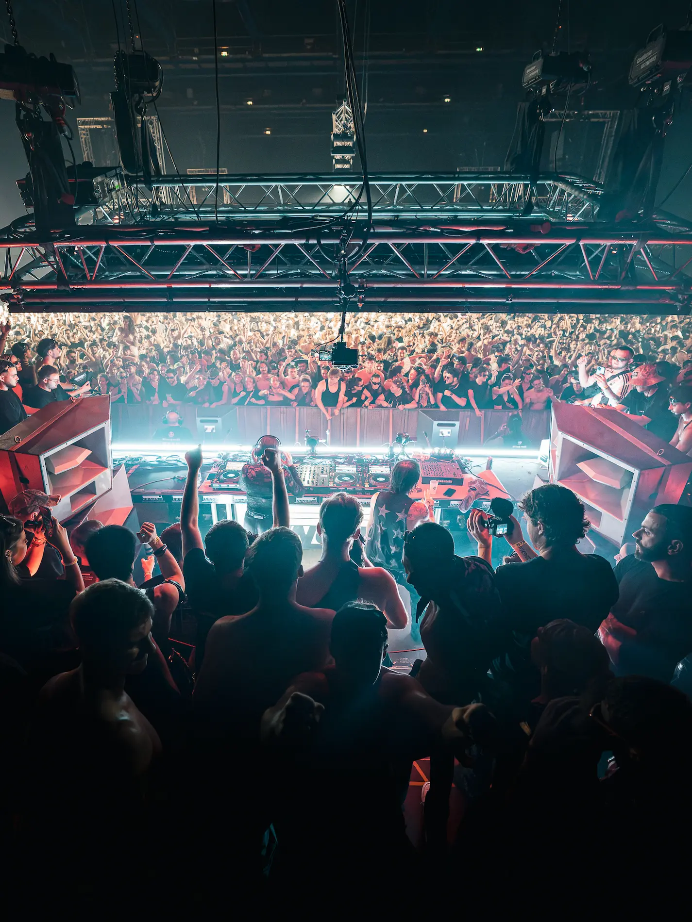 Un artiste en sweat à capuche bleu et bonnet rose chante sur scène devant une foule avec des téléphones levés sous un éclairage jaune vif.