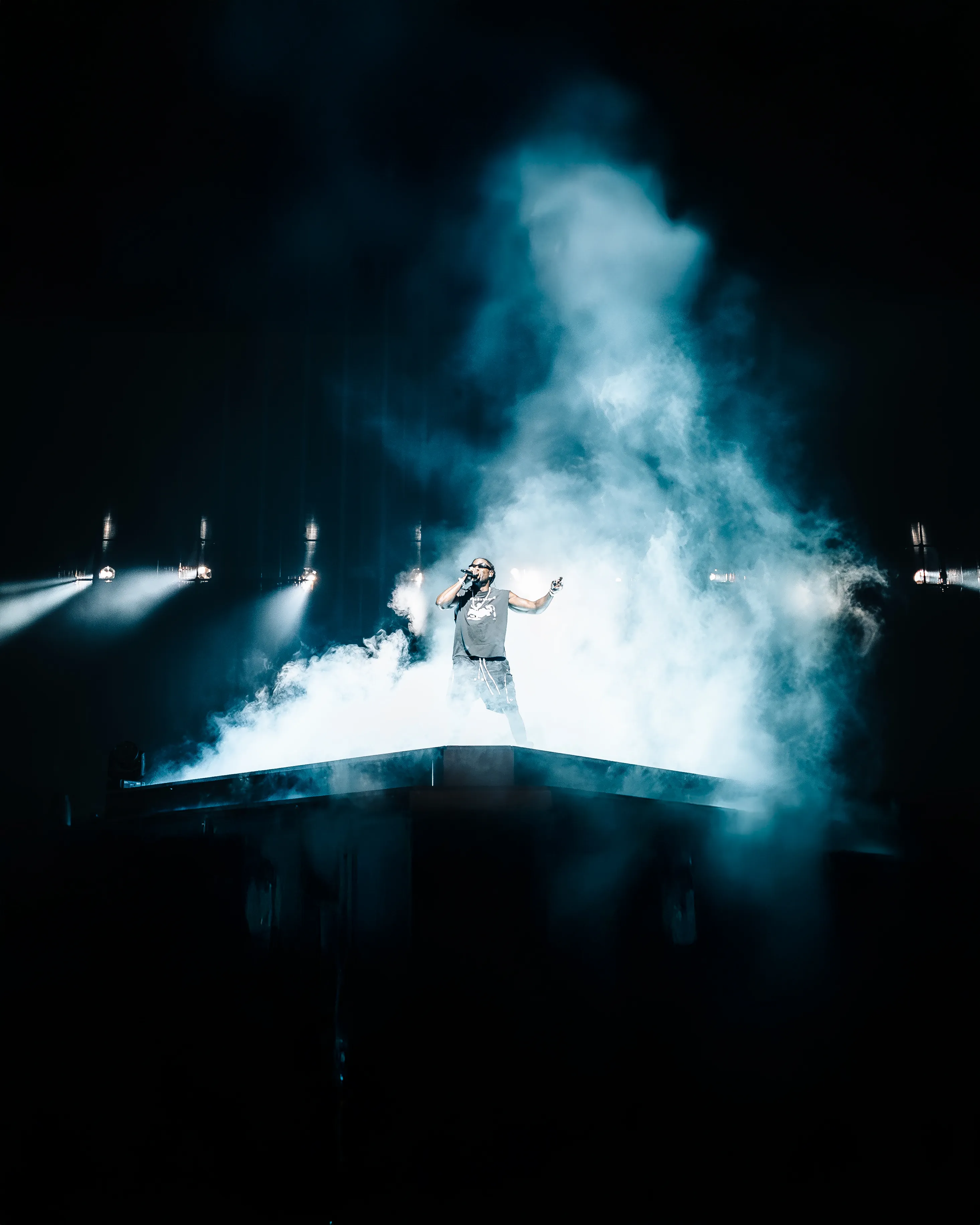 Artiste rap sur scène en sweat à capuche et bonnet, tenant un micro devant une foule avec des téléphones portables levés sous une lumière jaune intense.