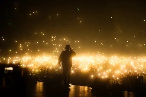 Un rappeur en sweat à capuche et bonnet rose tient un micro et se penche vers une foule illuminée par des téléphones portables levés lors d'un concert.