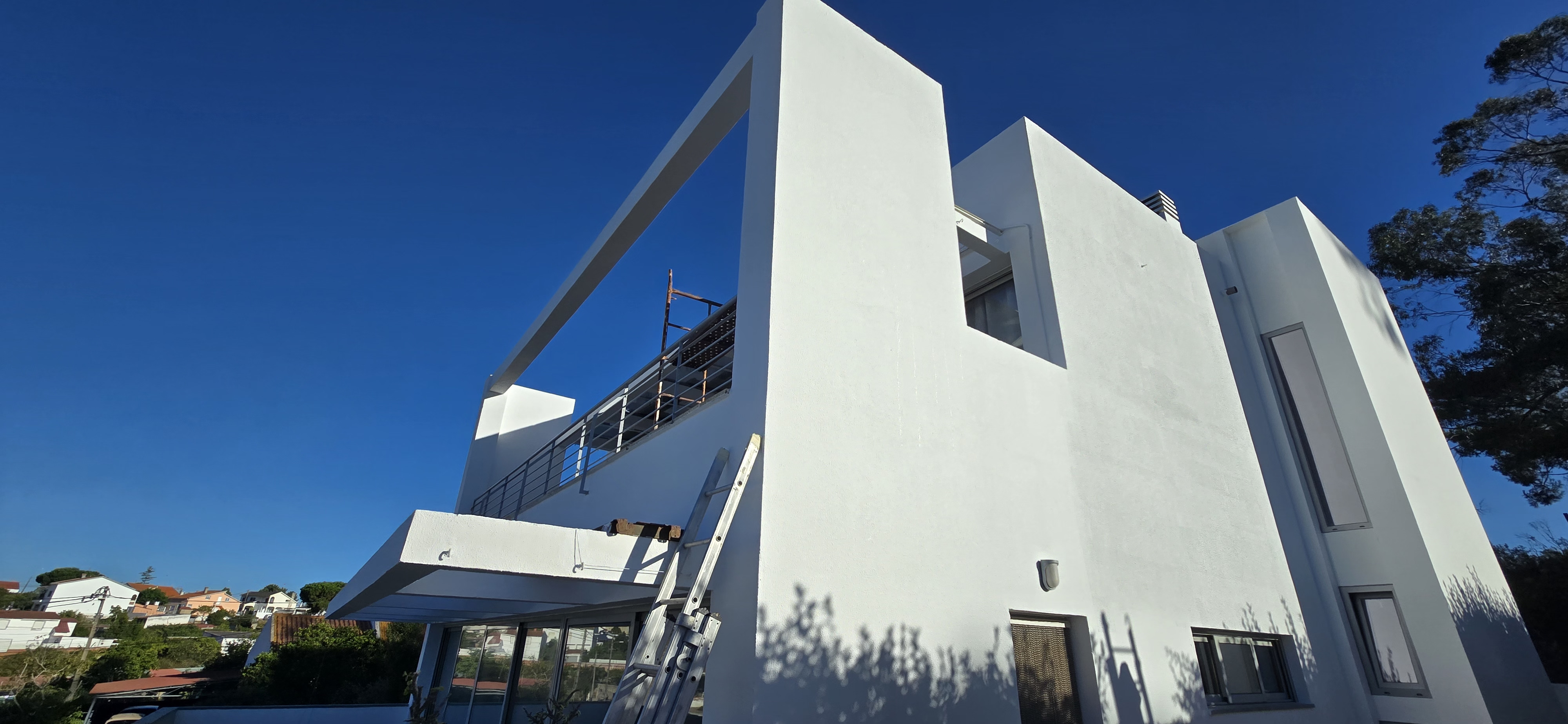 Casa branca moderna com janelas grandes, varanda com guarda-corpo metálico e escada encostada na parede.