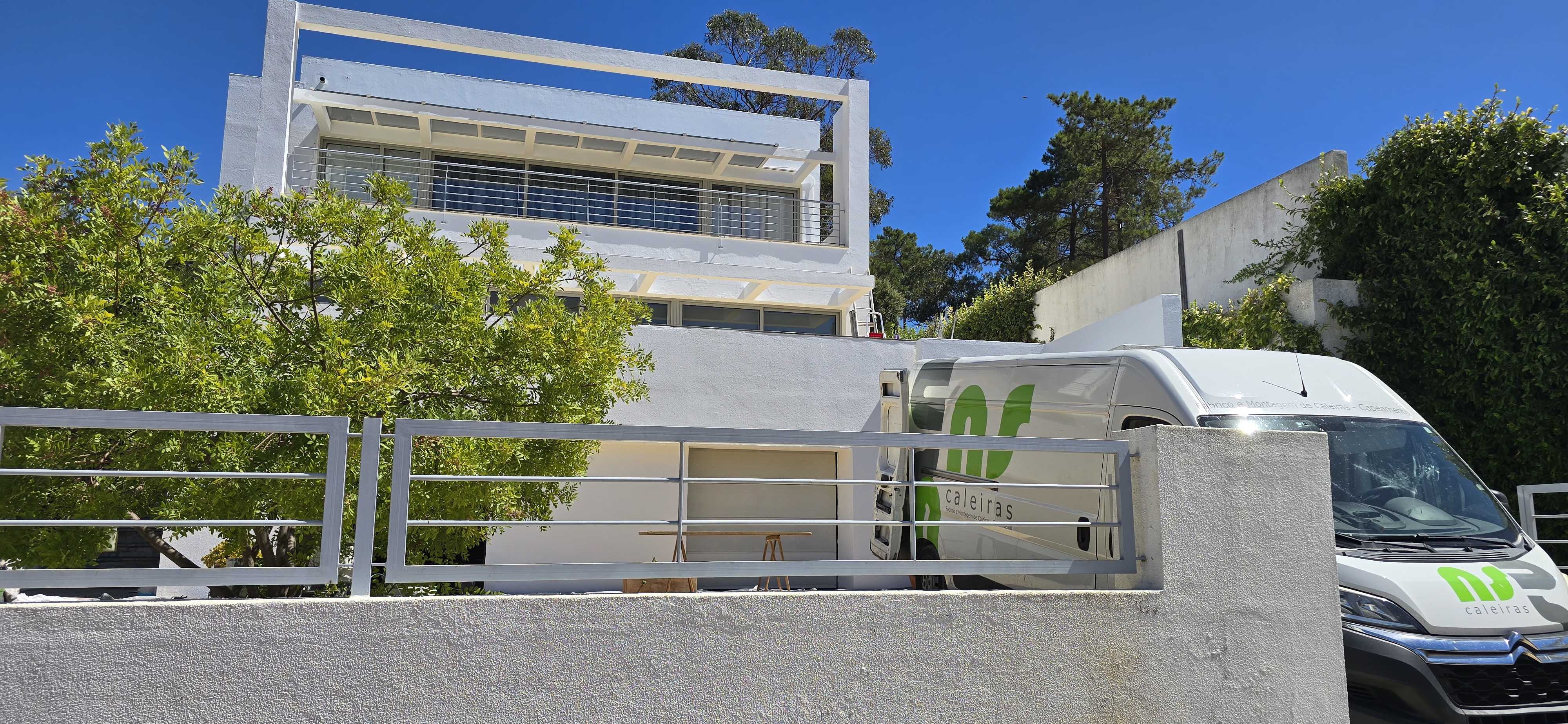 Fachada branca moderna com varanda, árvores verdes e carrinha estacionada com logo verde.