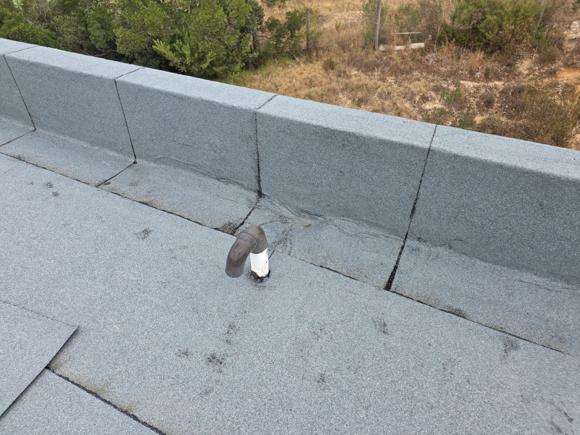 Cobertura de telhado cinzenta com tubo branco e preto e vegetação seca ao fundo.