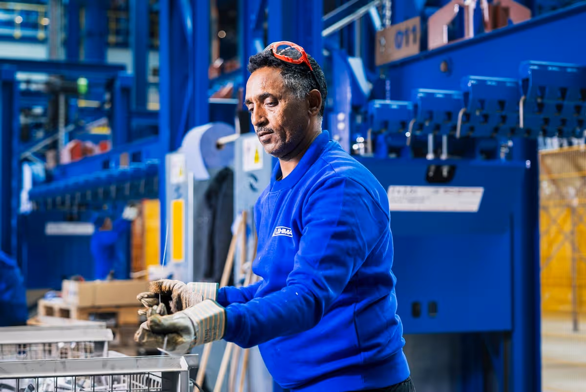 Ein Arbeiter in blauer Arbeitskleidung und Handschuhen, der in einer Fabrikhalle Metallteile inspiziert.