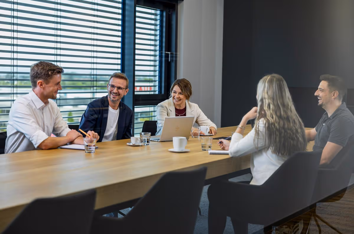 Fünf Personen in einem modernen Konferenzraum sitzen entspannt um einen langen Holztisch und führen eine freundliche Besprechung.