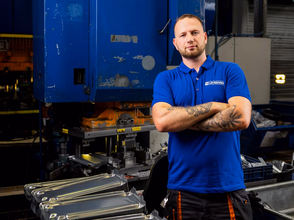 Mann mit Tätowierungen und blauem Lehmann-Poloshirt steht vor einer großen blauen Industriemaschine in einer Fabrikhalle.
