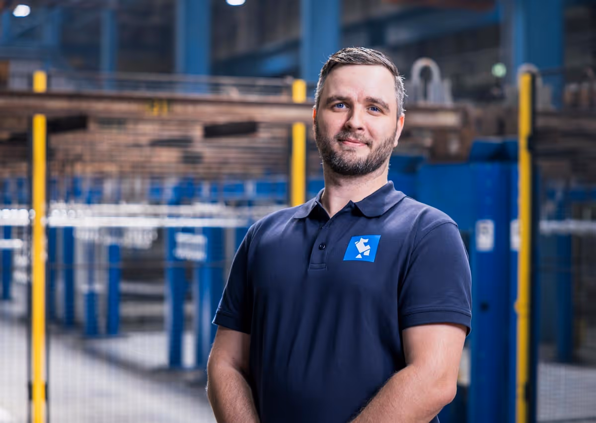 Ein Mann mit Bart trägt ein dunkelblaues Polohemd mit Logo und steht in einer industriellen Lagerhalle.