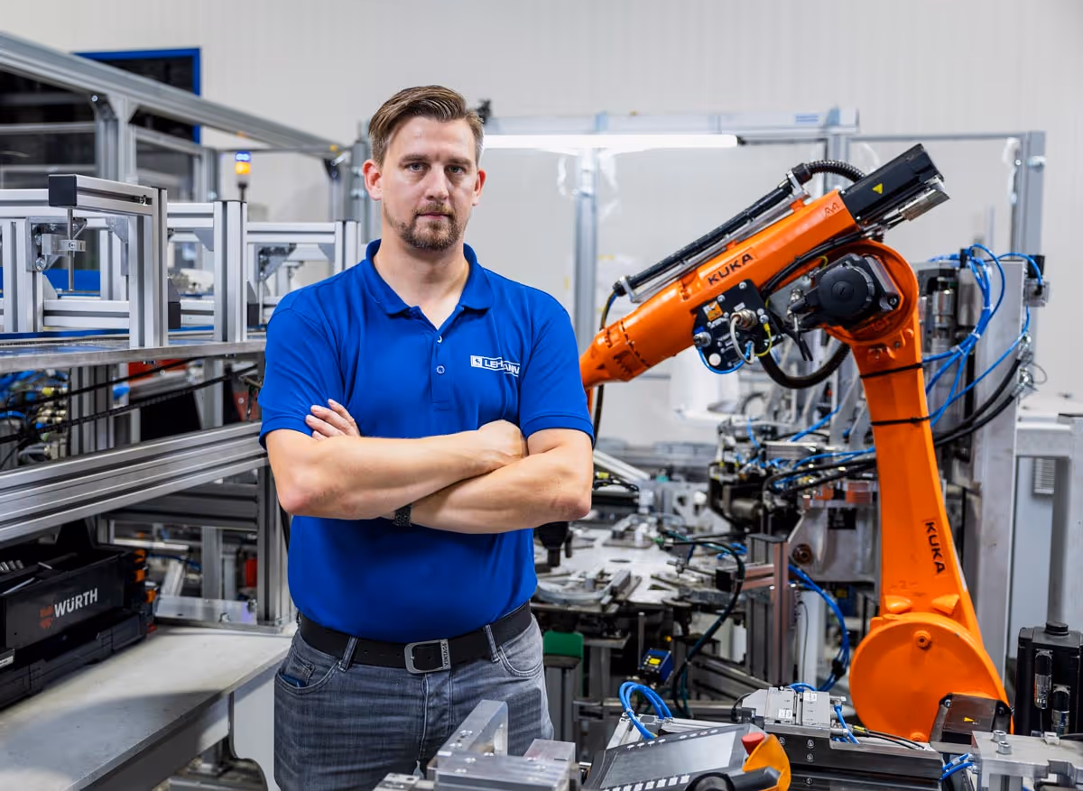 Mann in blauem Hemd steht mit verschränkten Armen in einer Fabrikhalle neben einem orangefarbenen Kuka-Industrieroboter.