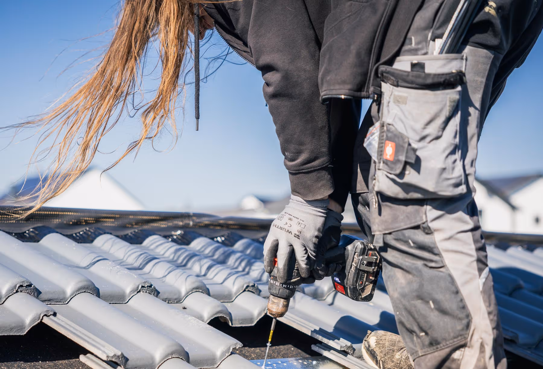 Handwerker mit langem Haar bohrt eine Schraube in graue Dachziegel auf einem Hausdach.