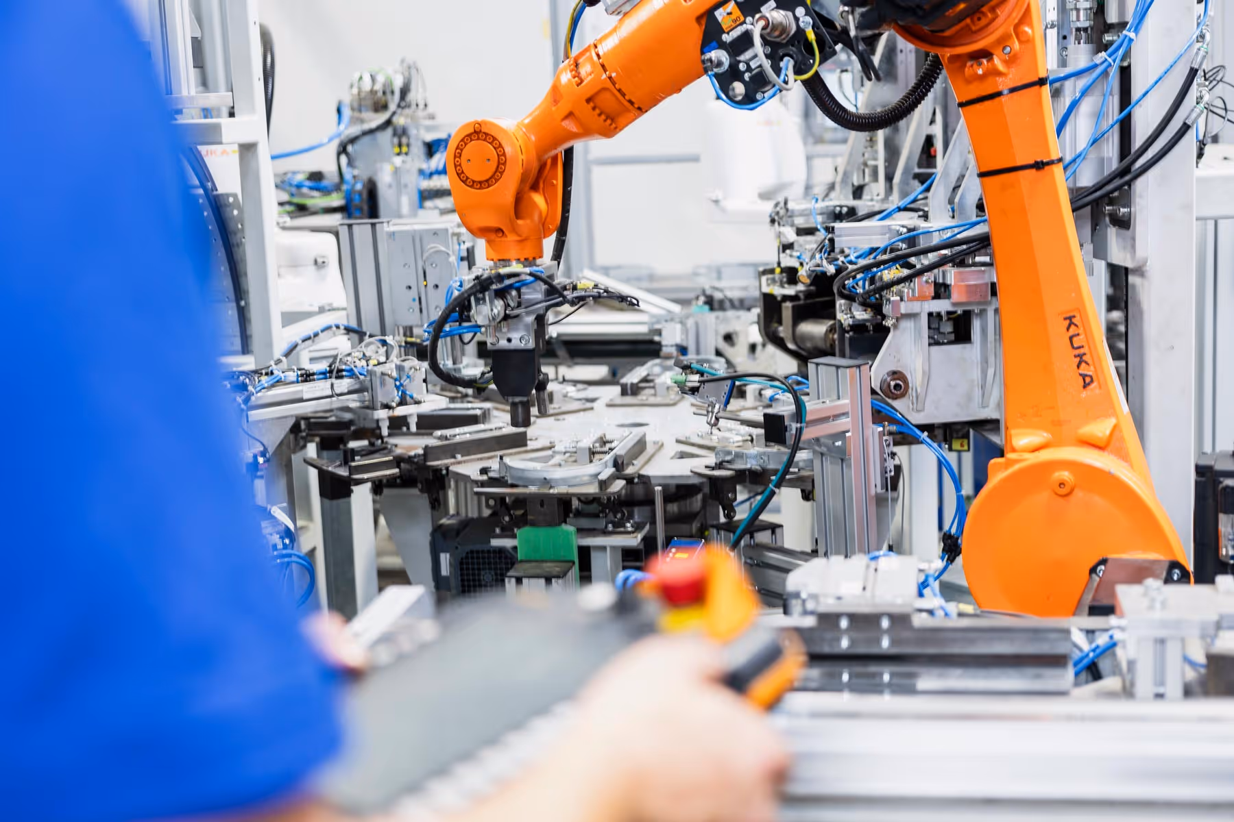 Industrieroboter arbeitet an einer Fertigungsmaschine, während eine Person mit einem Steuergerät im Vordergrund bedient.