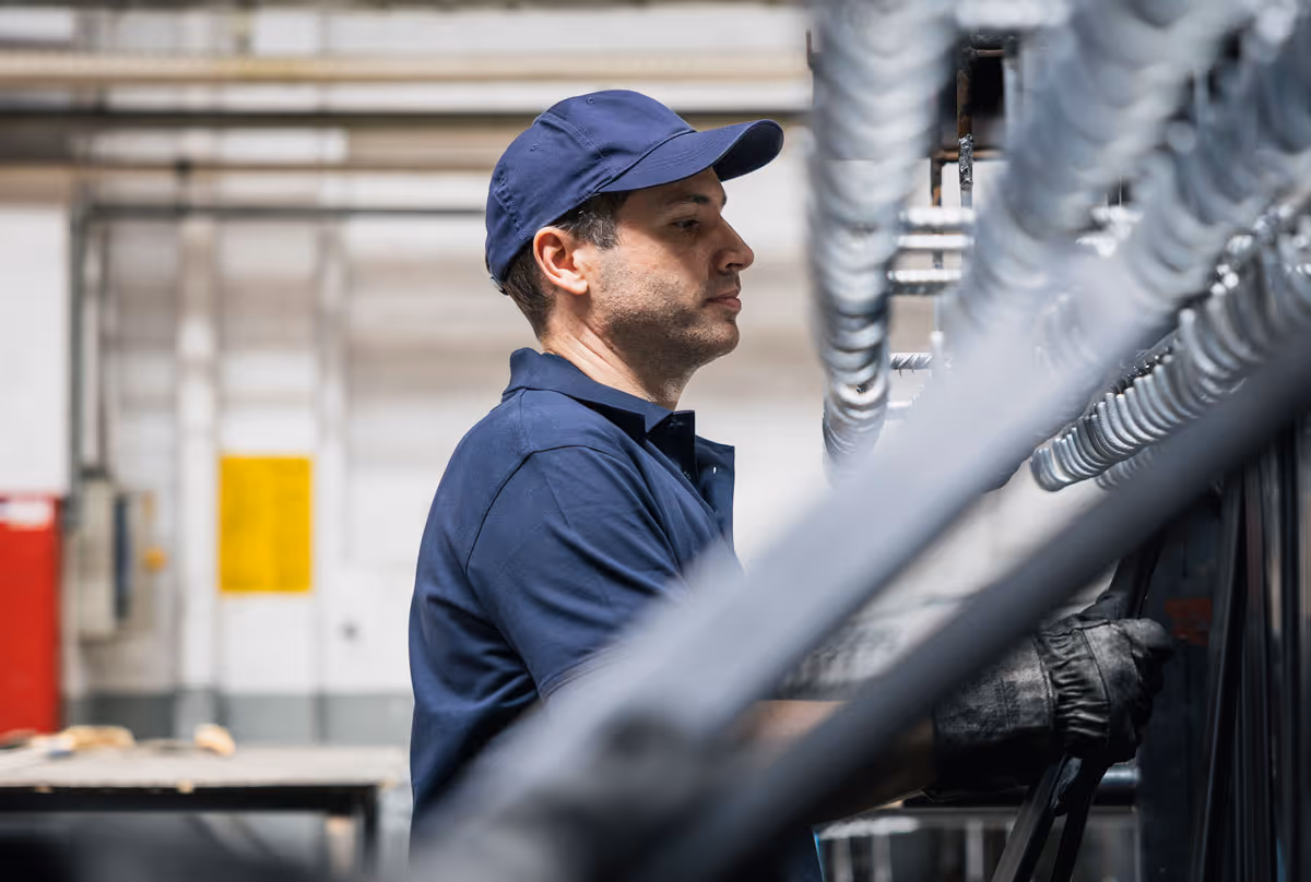 Ein Arbeiter in blauer Arbeitskleidung und Kappe bedient Maschinenteile in einer Fabrikumgebung.