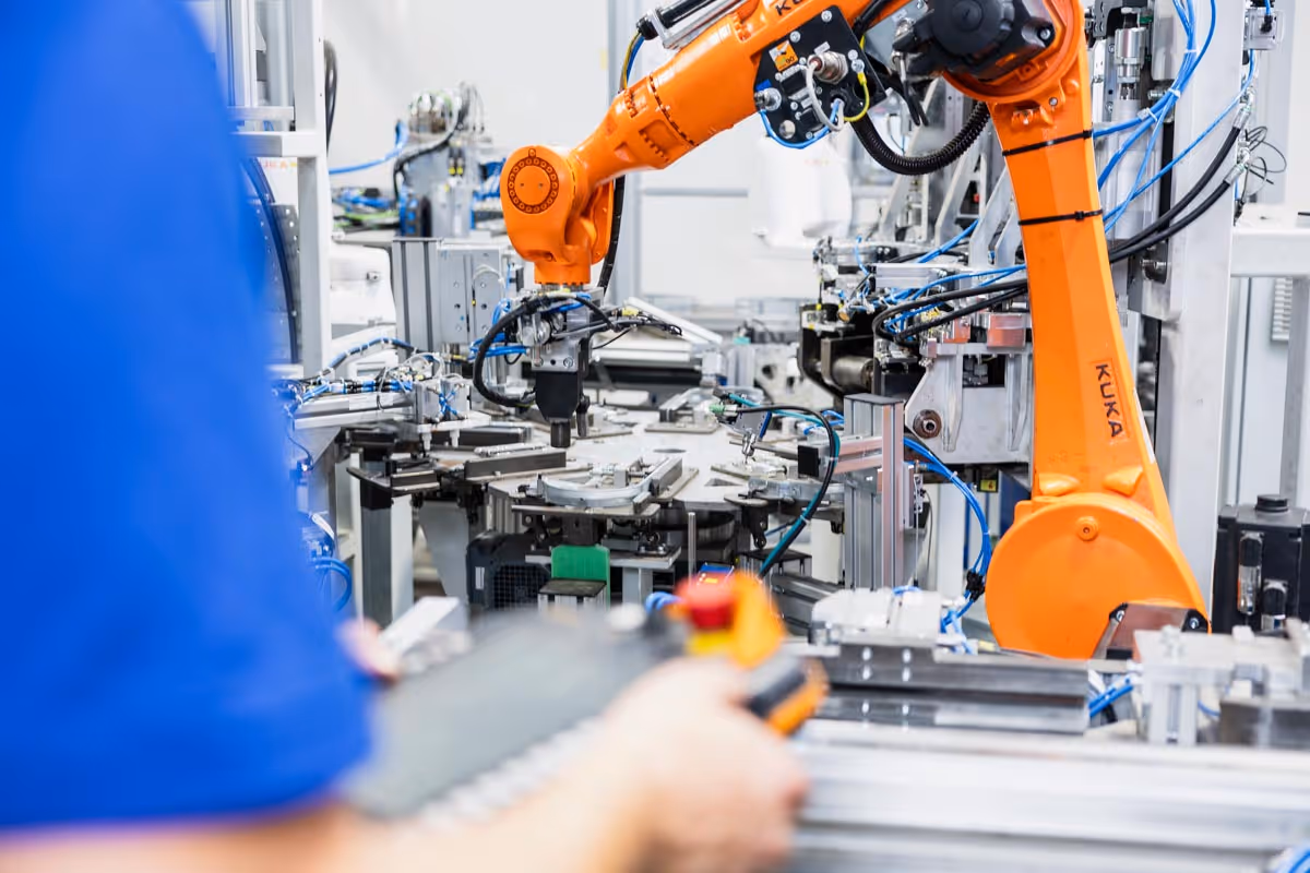 Industrieroboterarm steuert eine Maschine in einer Fabrik, während eine Person einen Bedienpult hält.