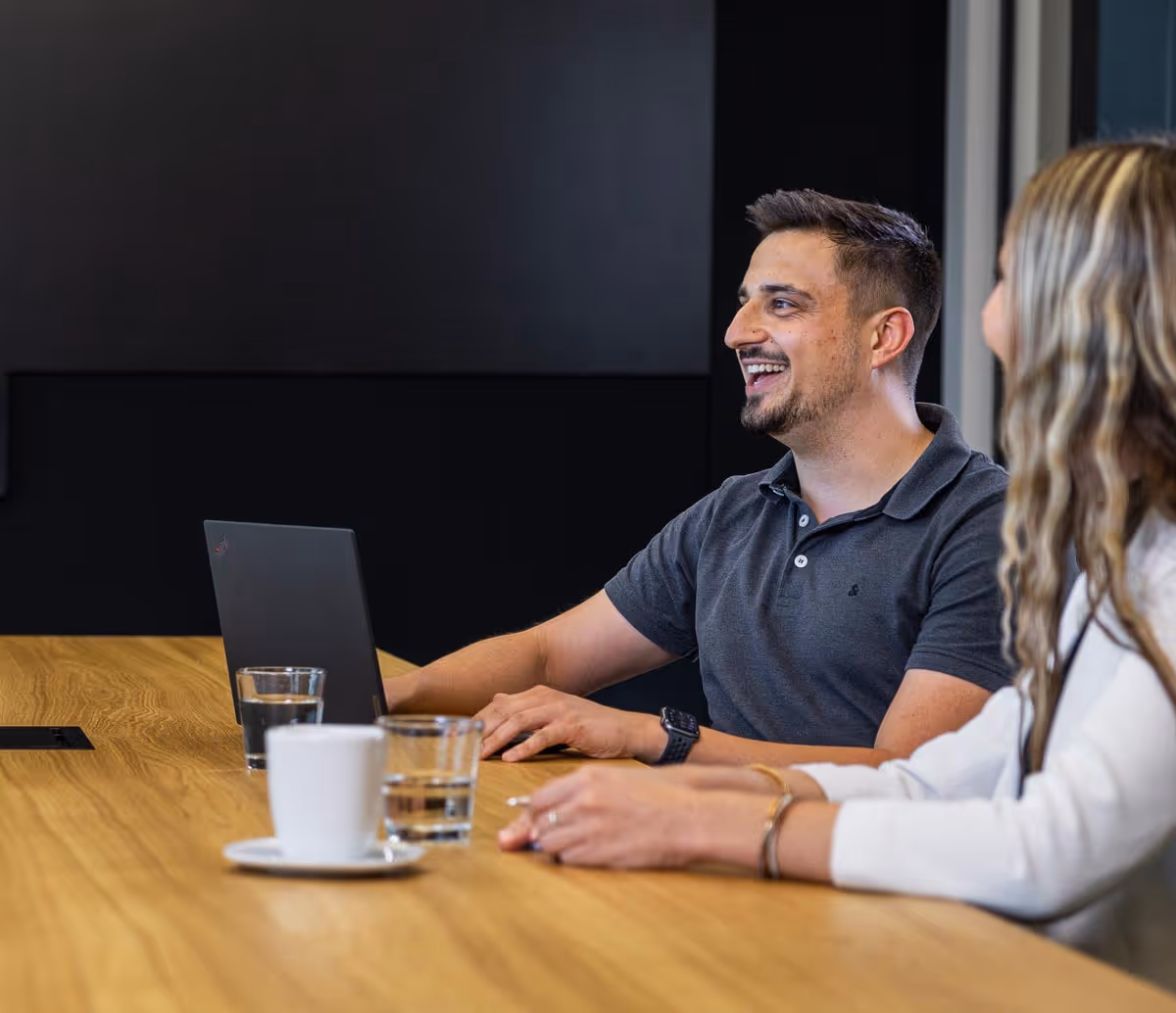 Zwei Personen sitzen an einem Holztisch, ein Mann mit Laptop lächelt, eine Frau ist im Vordergrund unscharf.