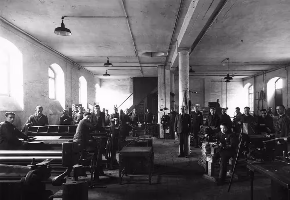 Schwarzweiß-Fabrikhalle mit zahlreichen Arbeitern an Maschinen aus der frühen Industriezeit.