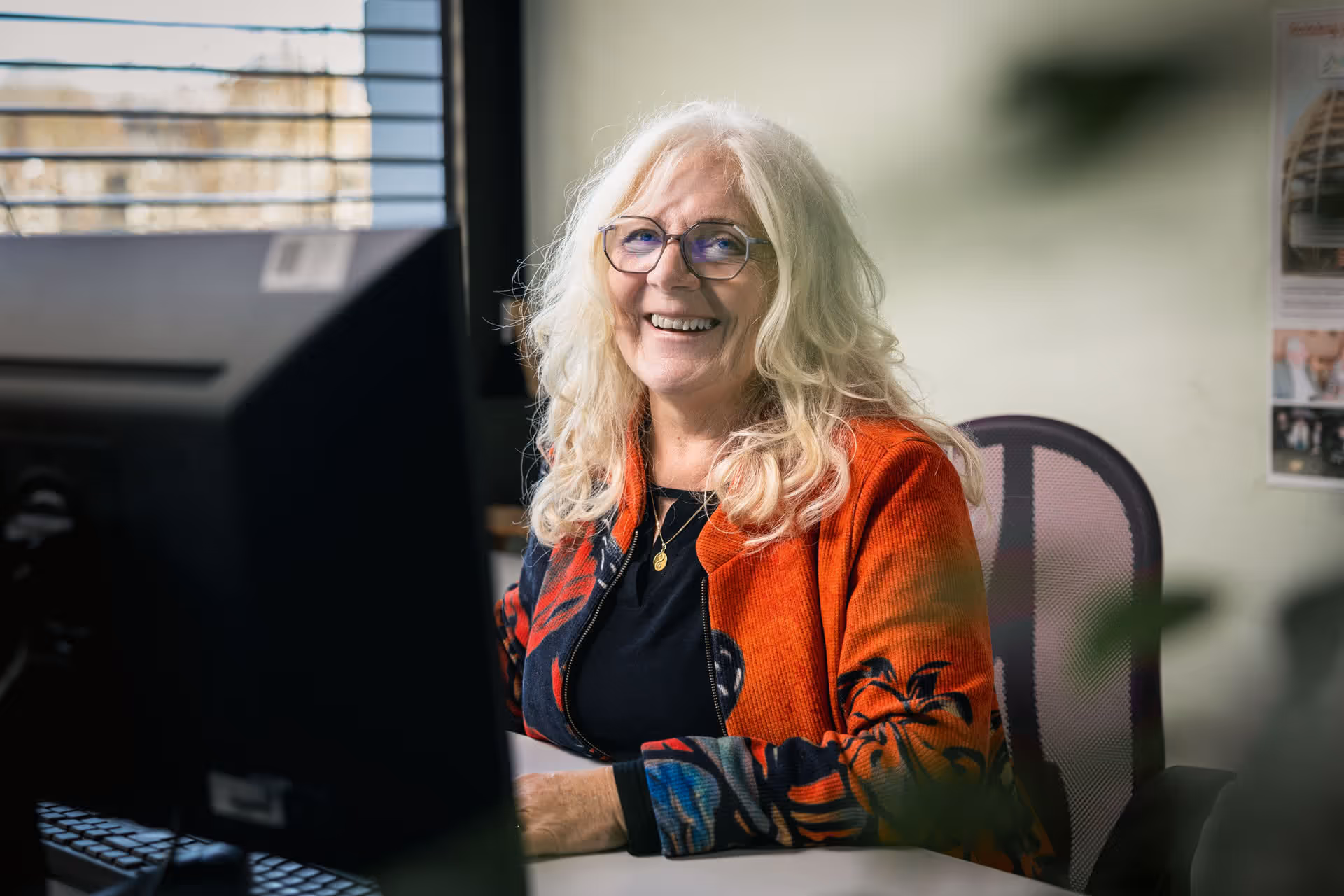 Lächelnde ältere Frau mit blonden Haaren und Brille sitzt am Schreibtisch vor Computer.