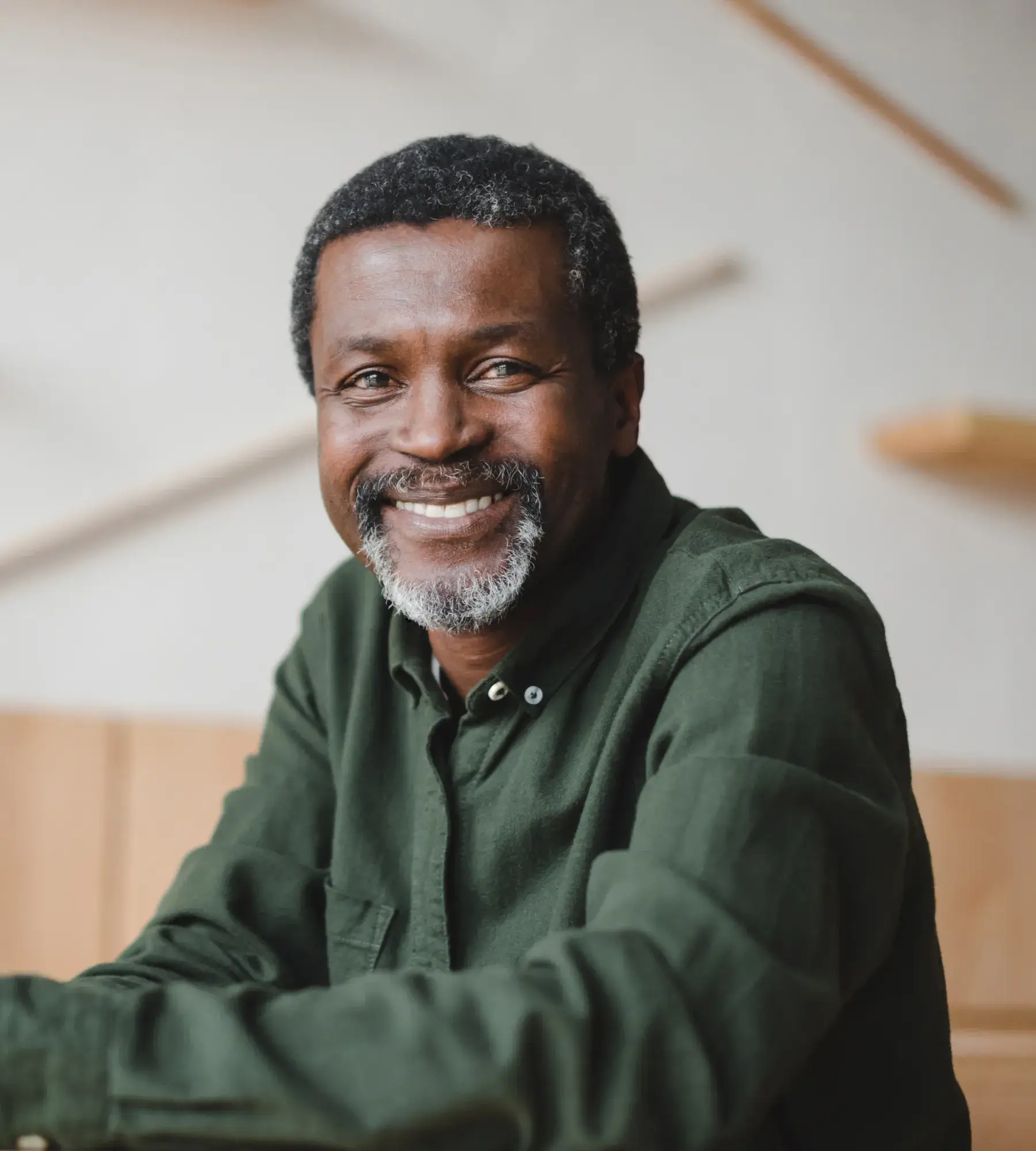 A man with a salt-and-pepper beard smiles warmly while sitting indoors, wearing a dark green shirt.