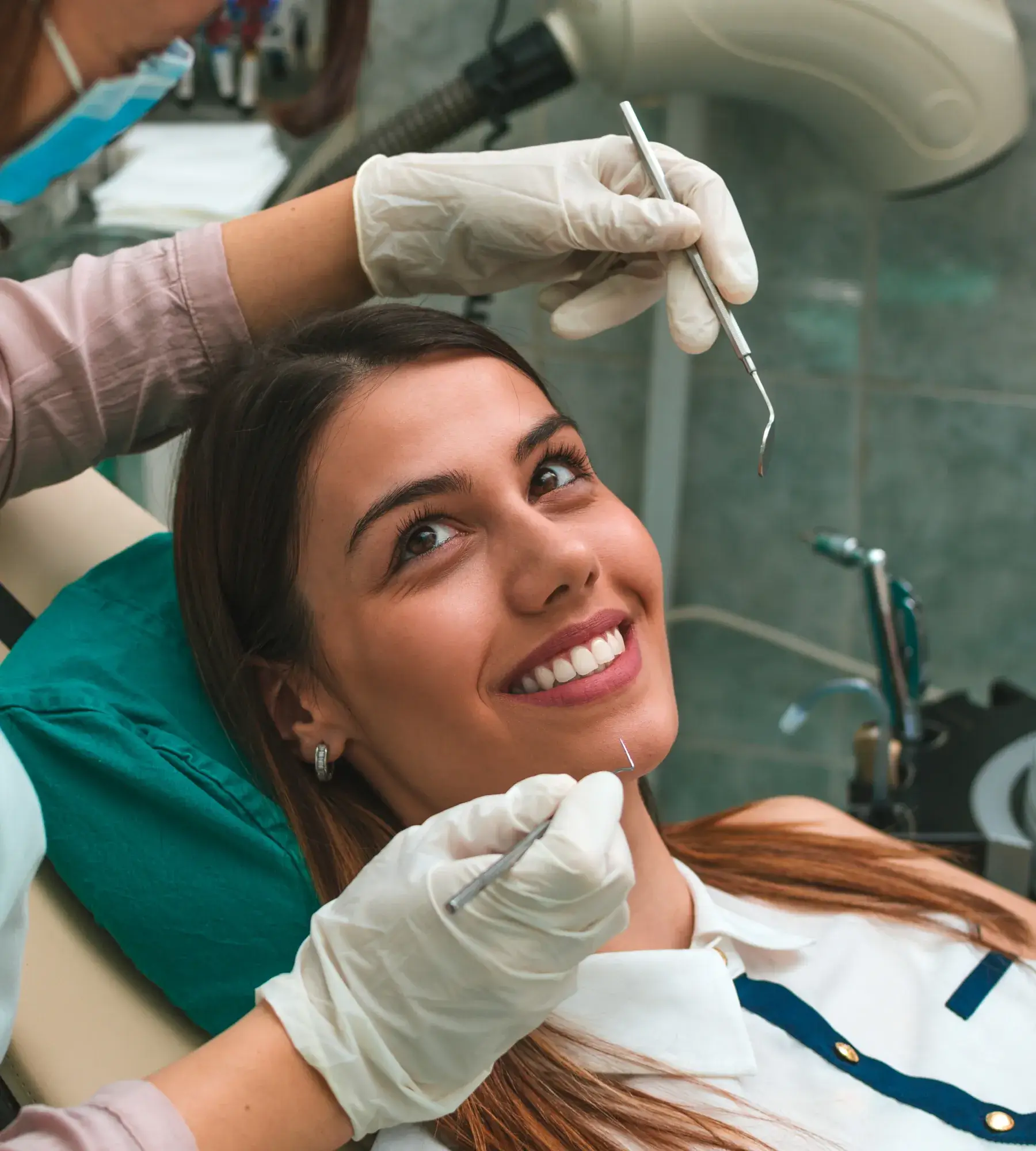 A woman smiles while sitting in a dentist's chair, as a dentist prepares dental tools.