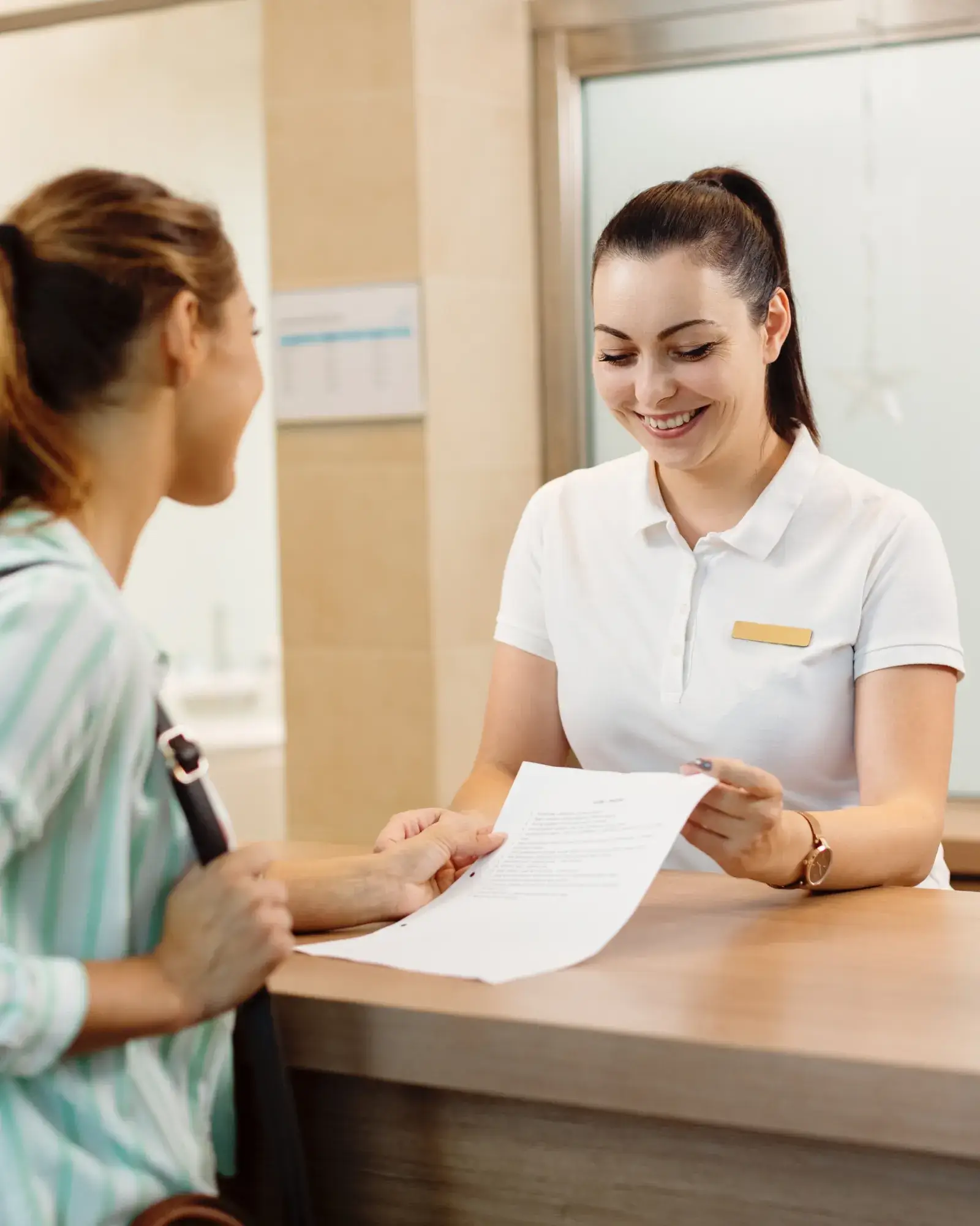 A receptionist hands a document to a woman across the counter, both smiling.