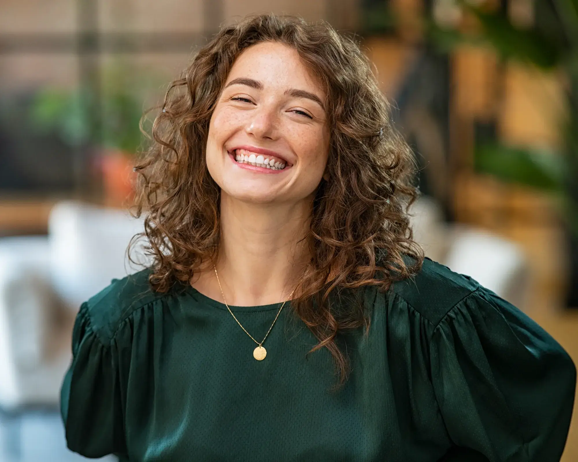 A person with curly hair smiles brightly, wearing a green blouse and a gold necklace.