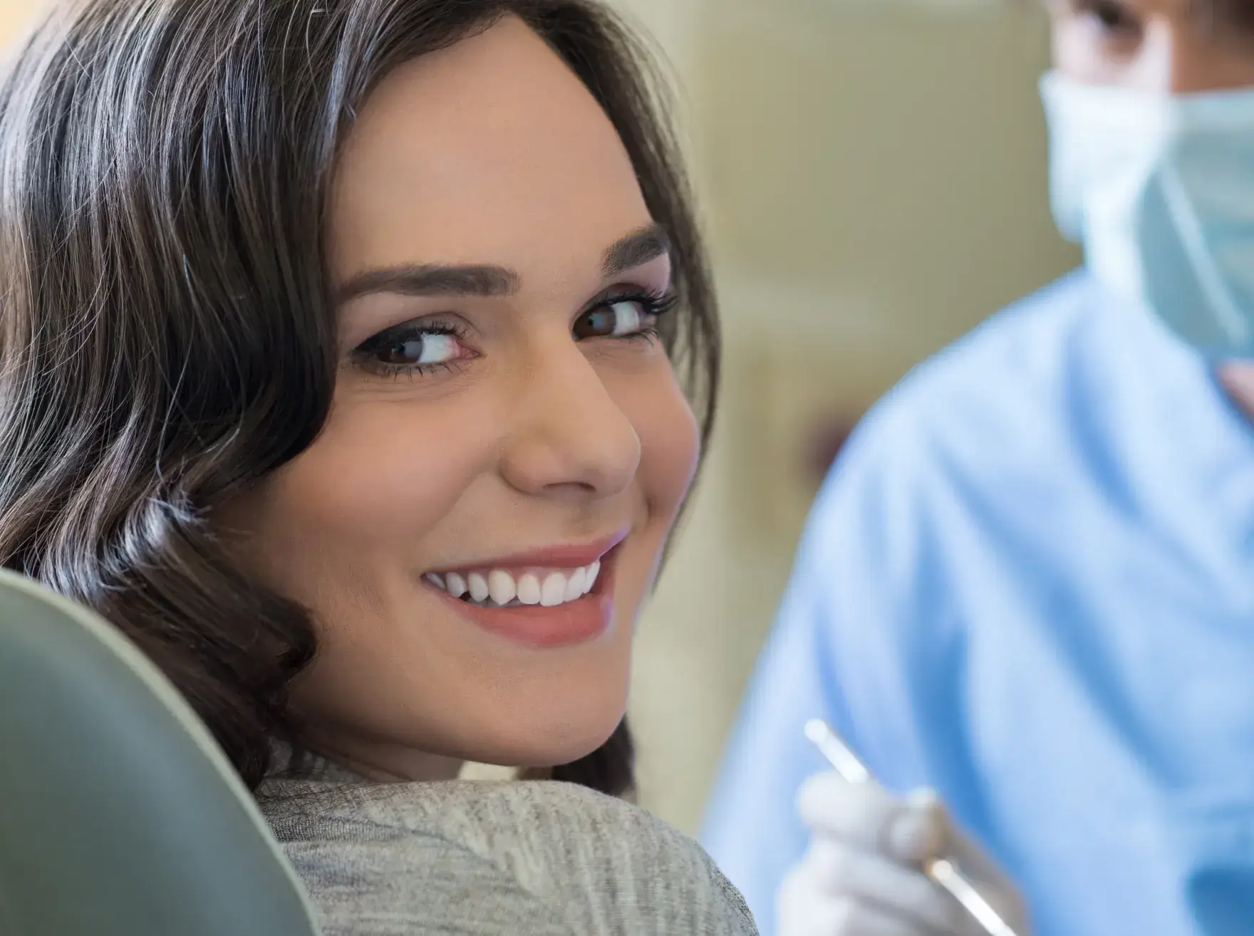 Woman smiling while sitting in a dentist's chair, with a dental professional beside her.